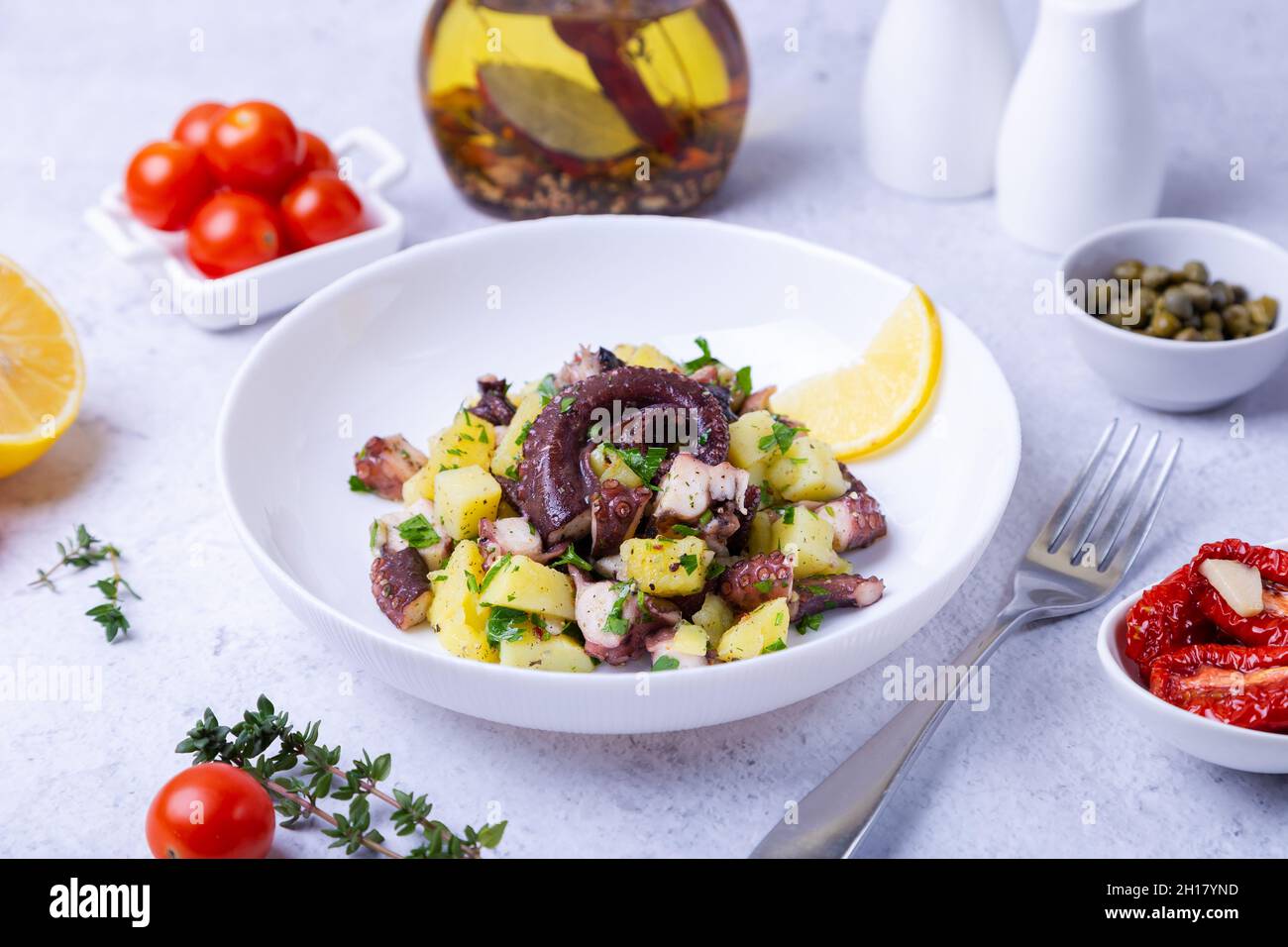 Warm salad with octopus, potatoes, tomatoes, capers and lemon on a white plate. Close-up, white background. Stock Photo