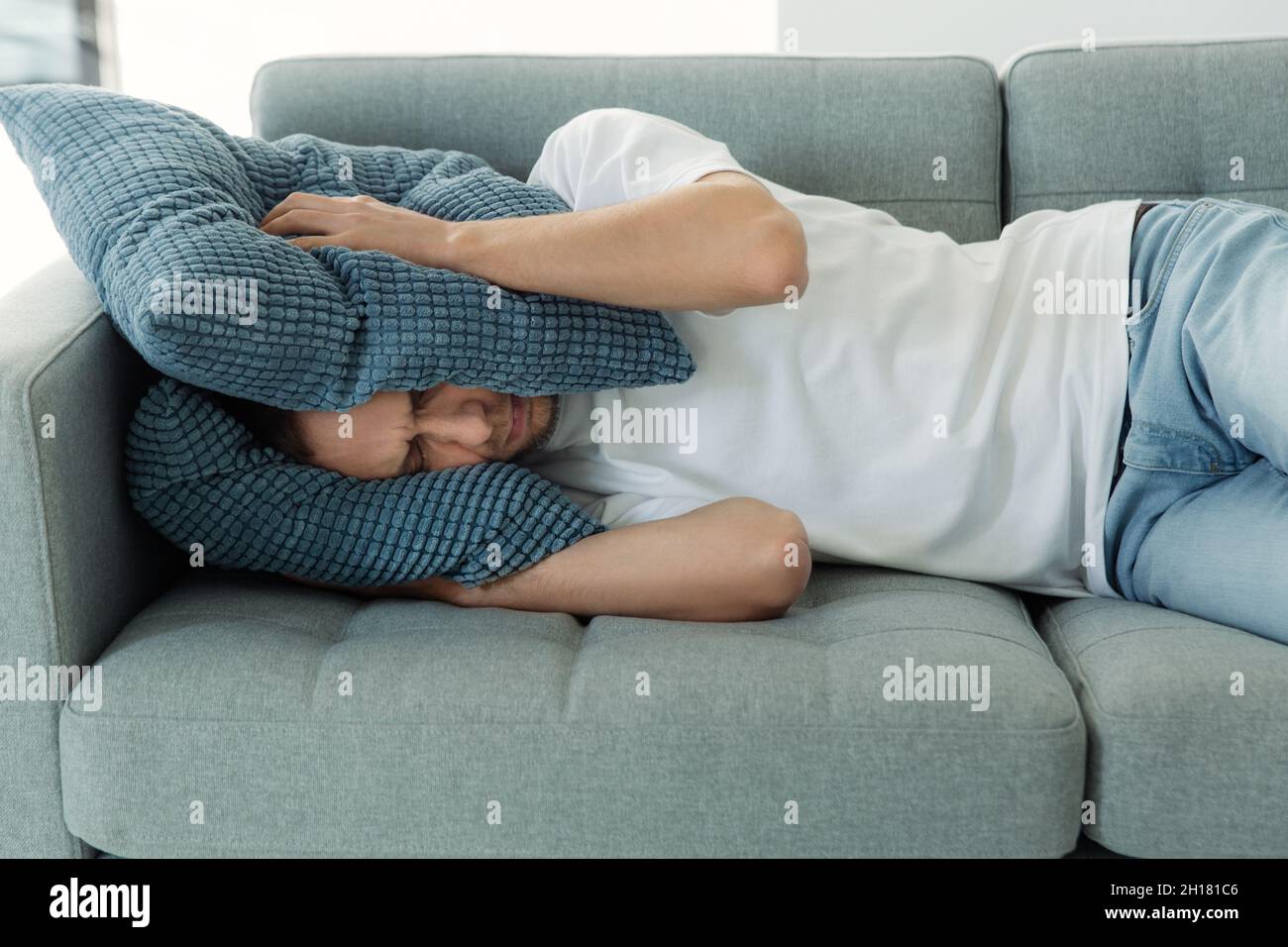 Sleepless man tired of noisy sounds from neighbors try to sleep on sofa covering head with pillows Stock Photo