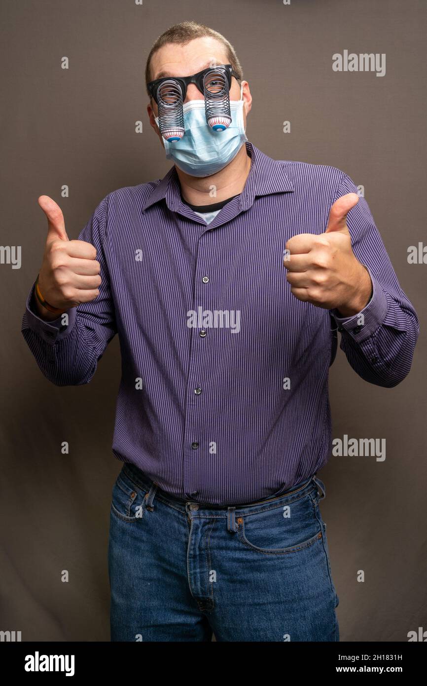 Male wearing a medical facemask and funny droopy eyes glasses standing with a thumbs-up gesture Stock Photo