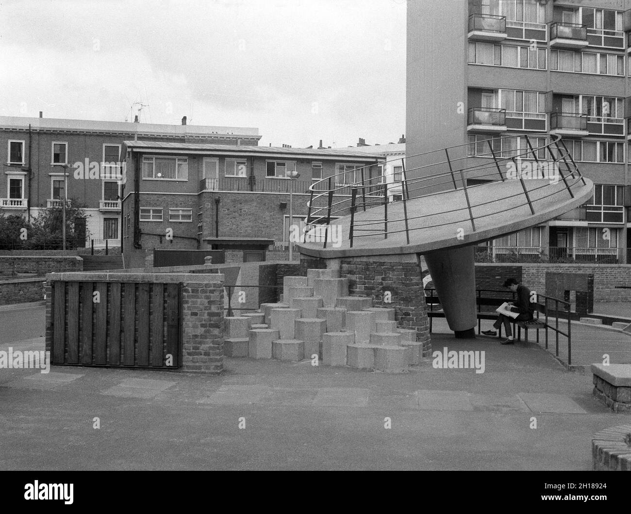 1960s, historical, a concrete made playground area outside a large council housing estate, Churchill Gardens in Pimlico, inner London, England, UK. Built between 1948 and 1962 to replace victorian terraces damaged in the Blitz of WW2, this was a huge estate of social housing, with 32 tower blocks providing 1,600 homes. Seen in the picture a young man sitting on a bench underneath a concrete made playground saucer with railings, with hexagon steps up. Stock Photo