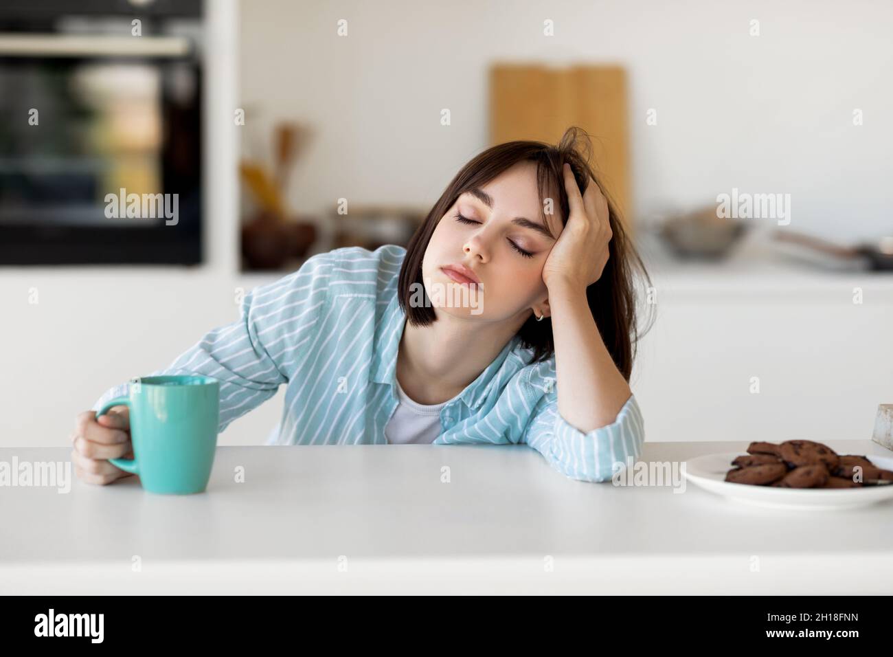 Sleepy young woman drinking coffee, feeling tired, suffering from insomnia and sleeping disorder, sitting in kitchen Stock Photo