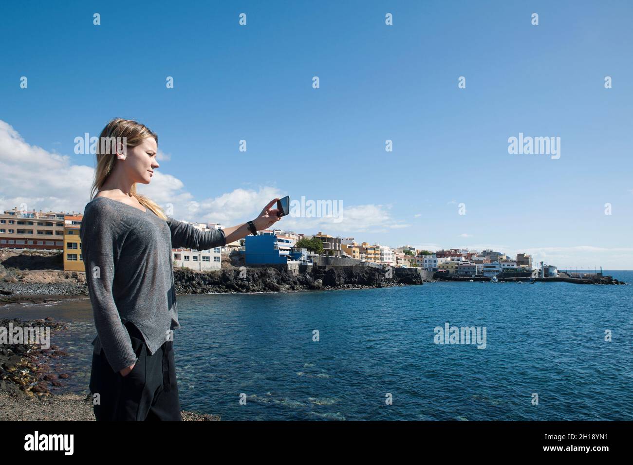 Beautiful millennial woman holding a smart phone and taking a selfie or a photo of the views of the fishing village Los Abrigos, Tenerife Stock Photo