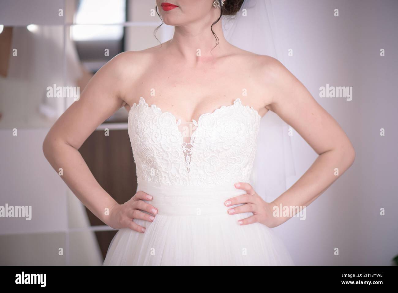 Cropped semi-profile shot of a Caucasian woman wearing a red hot lipstick and a bridal sweetheart neckline dress while holding hands on the waistline Stock Photo