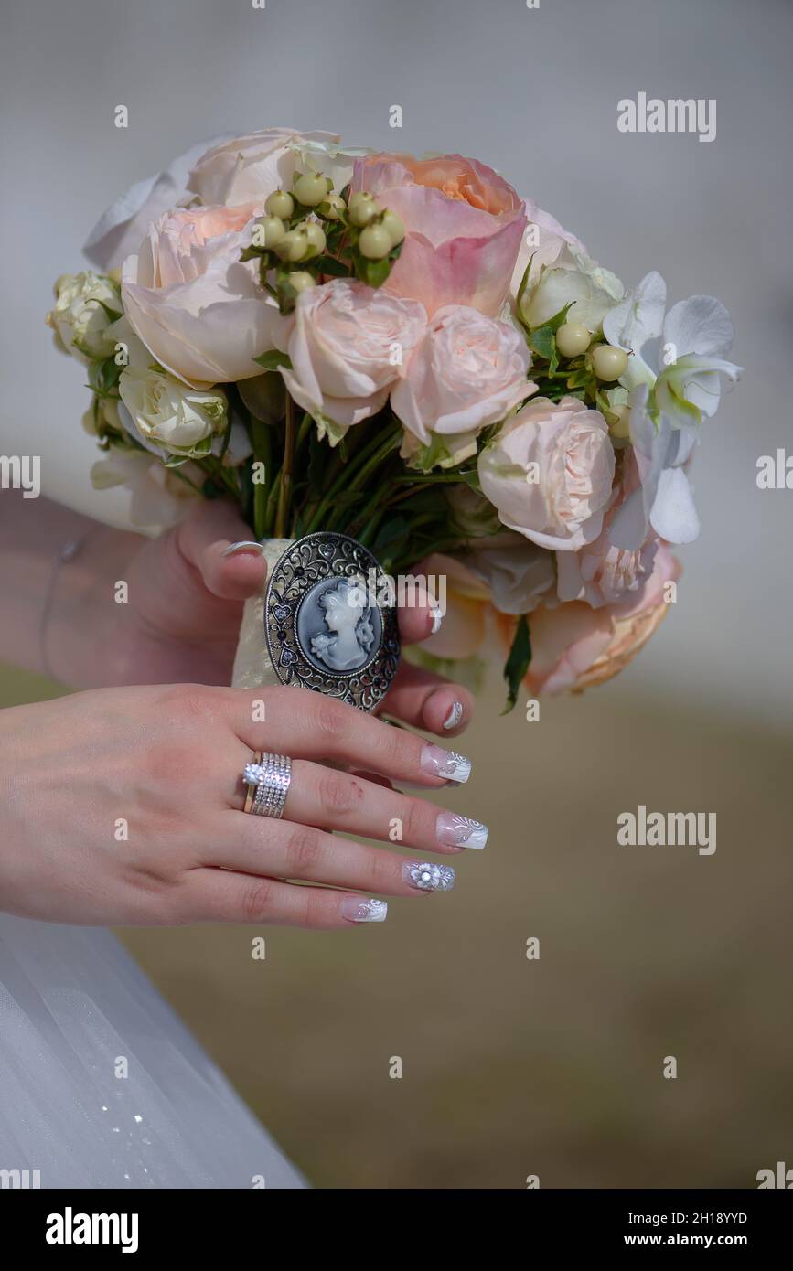 Close-up of Caucasian bride with impeccable manicure holding a round pastel bouquet featuring a vintage cameo brooch with large white orchids Stock Photo
