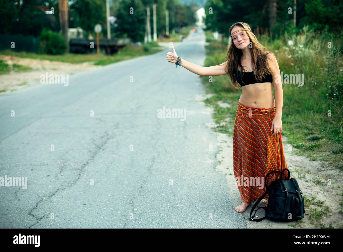 A hippie girl voting near the rural road. Hitchhiking trips. Stock Photo