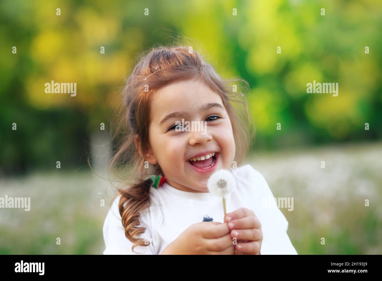 Pretty  little girl  is smiling in the park with dandelion flower. Happy cute kid having fun outdoors at sunset. Stock Photo