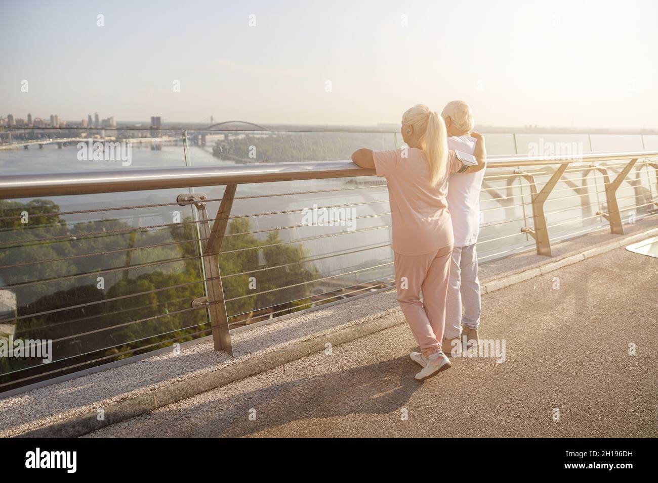 Senior lady hugs husband in sports clothes enjoying city view from footbridge Stock Photo