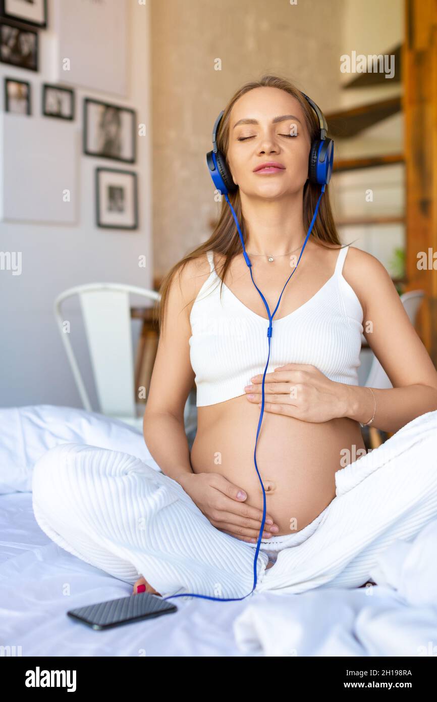A young pregnant woman sits on a bed wearing blue headphones and listens to soothing music through an online smartphone application. Stock Photo