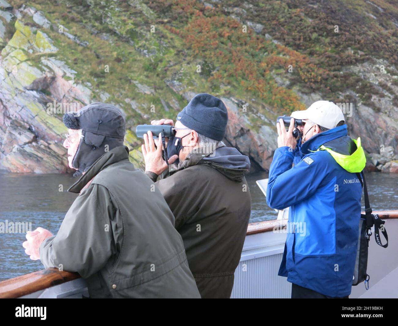 Three men, two with binoculars, at the stern of a boat, bird spotting as a cruise ship sails through the majestic landscape of Loch Torridon. Stock Photo