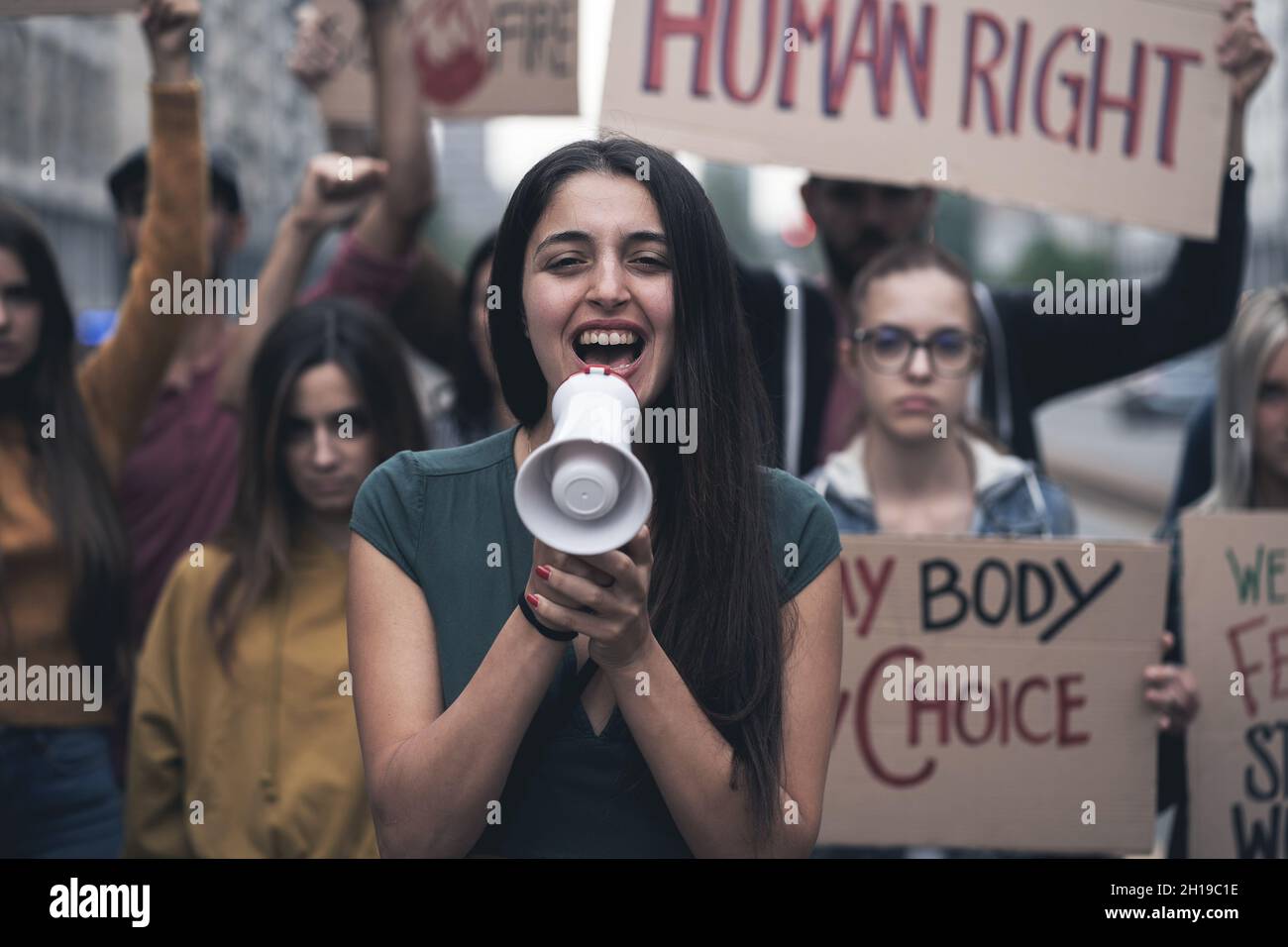 Group of young people protesting on the street with signs raised to promote human rights. Focus on the woman in front screaming to the megaphone Stock Photo