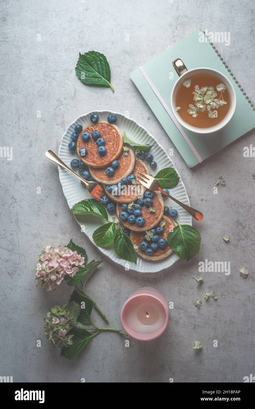 Blueberry pancakes breakfast on grey concrete kitchen table with petals, leaves, forks, tea, candle and notebook. Morning routine with healthy food an Stock Photo
