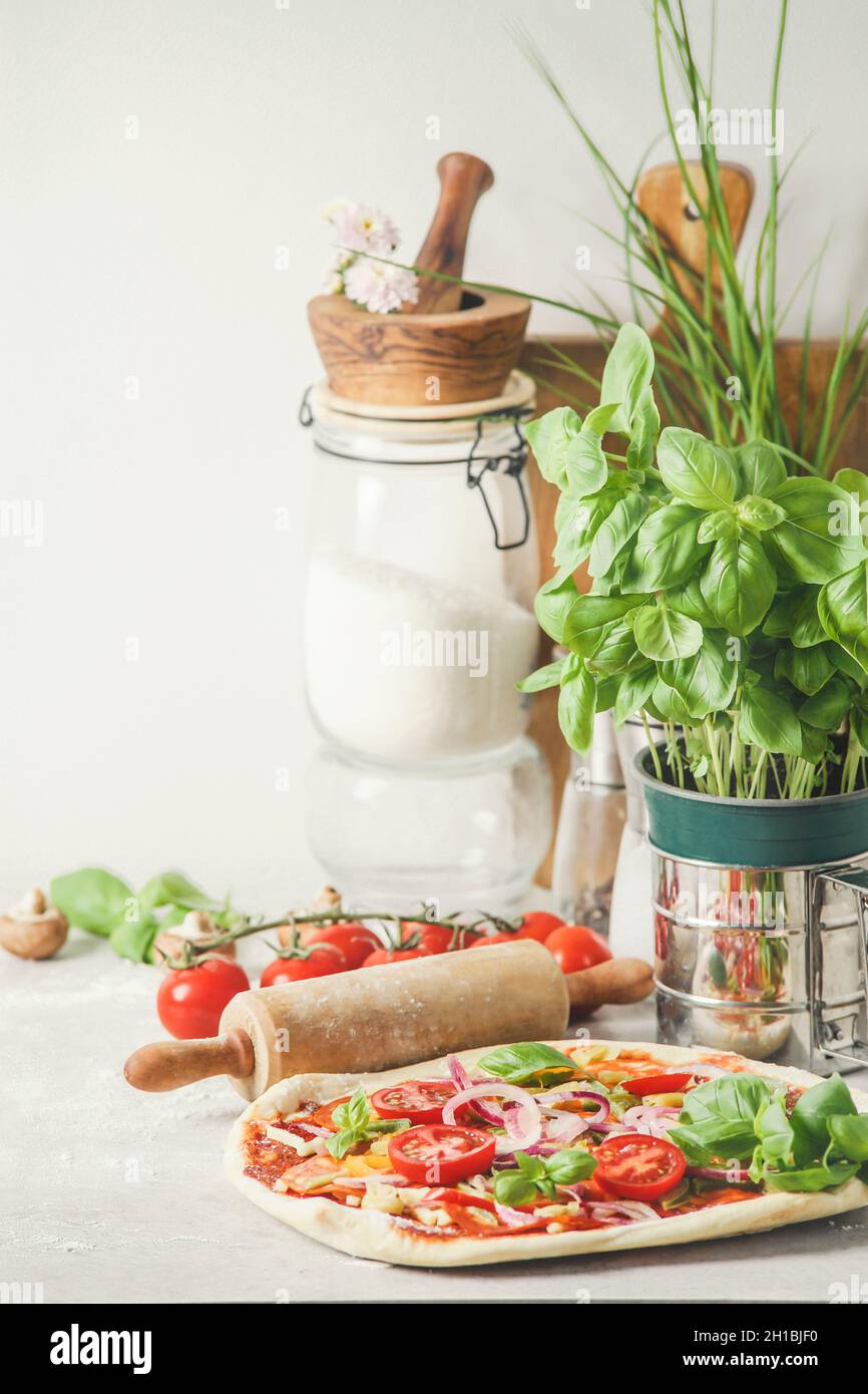 Vegan pizza with vegetables, basil, rolling pin, mortar and pestle, herbs, tomatoes and mushrooms at white kitchen background. Fresh cooking at home w Stock Photo