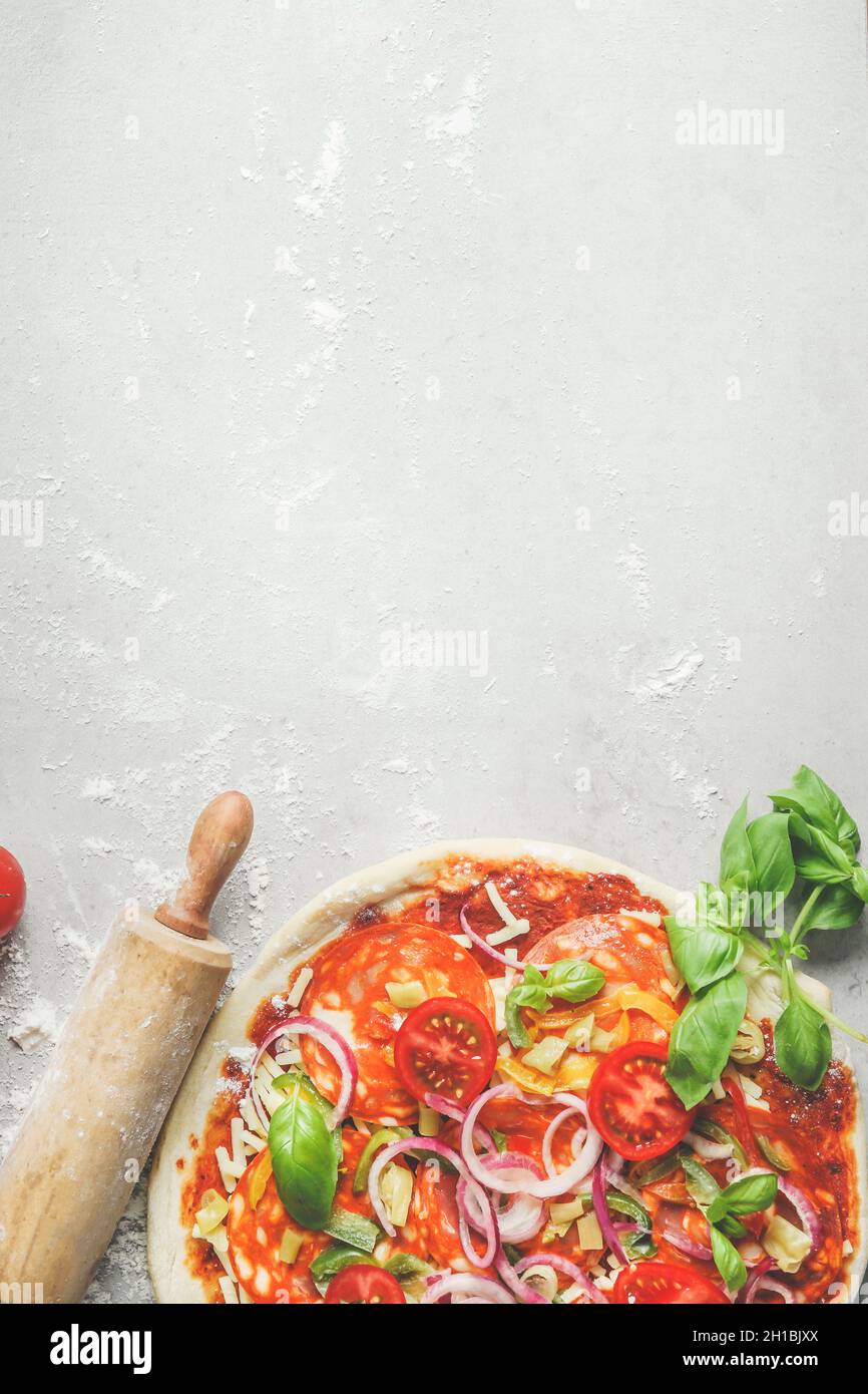 Homemade pizza background salami and tomatoes, wooden rolling pin, basil leaves on grey kitchen table with flour. Cooking italian food at home. Top vi Stock Photo
