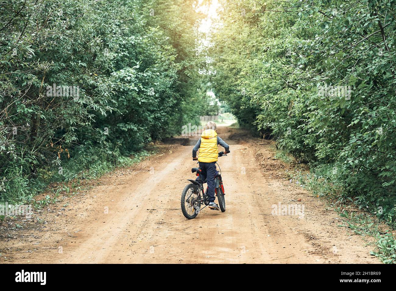 Little boy in yellow vest rides on modern bicycle on rural ground road with green trees on sides in summer Stock Photo