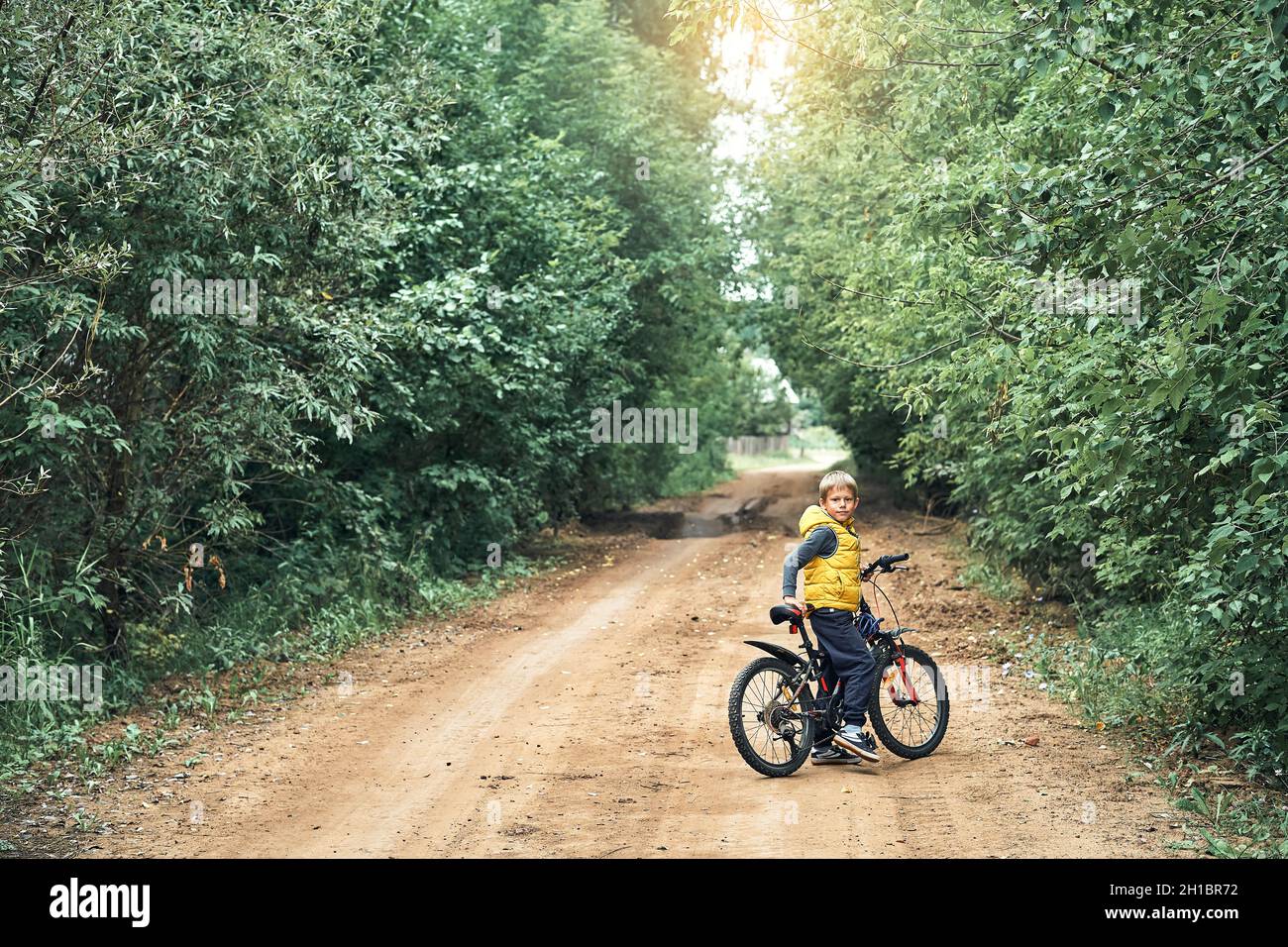 Adorable little boy in yellow vest with modern bicycle looks at camera on rural ground road with green trees on sides in summer Stock Photo
