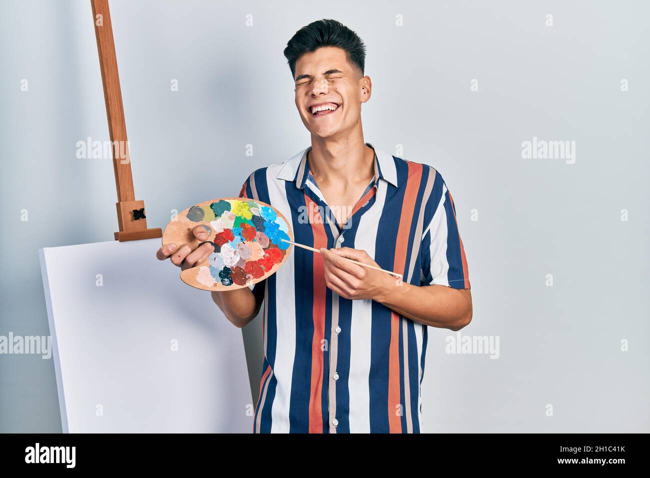 Young hispanic man holding painter palette and paintbrush standing