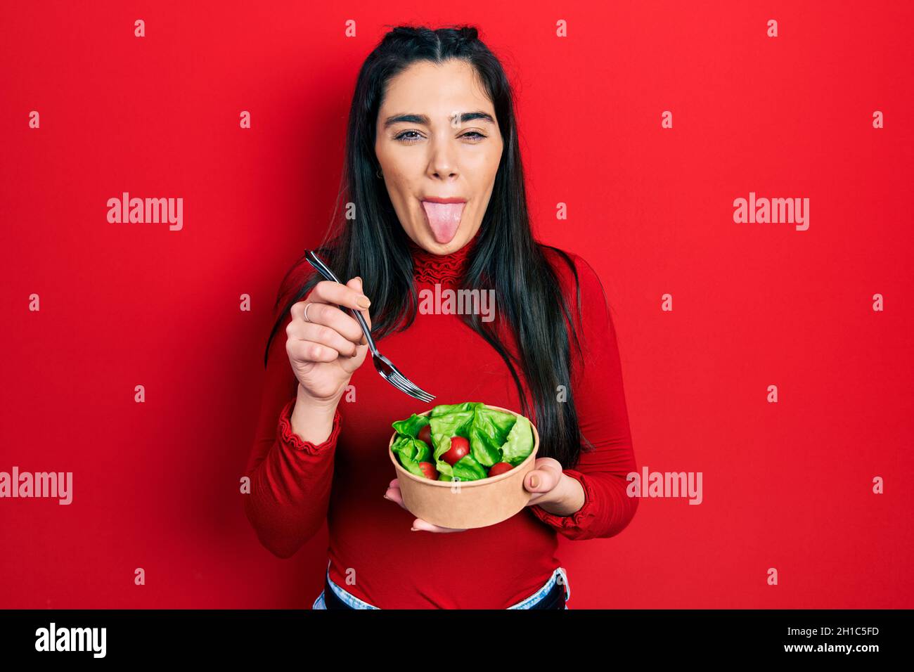 Young hispanic girl eating salad sticking tongue out happy with funny expression. Stock Photo