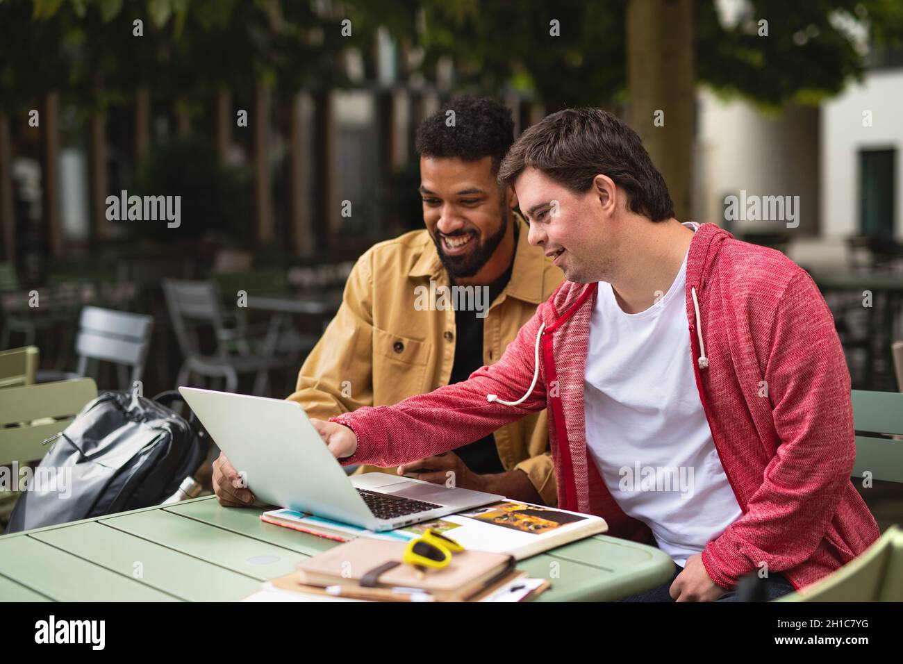 Young man with Down syndrome with his mentoring friend sitting outdoors in cafe using laptop. Stock Photo