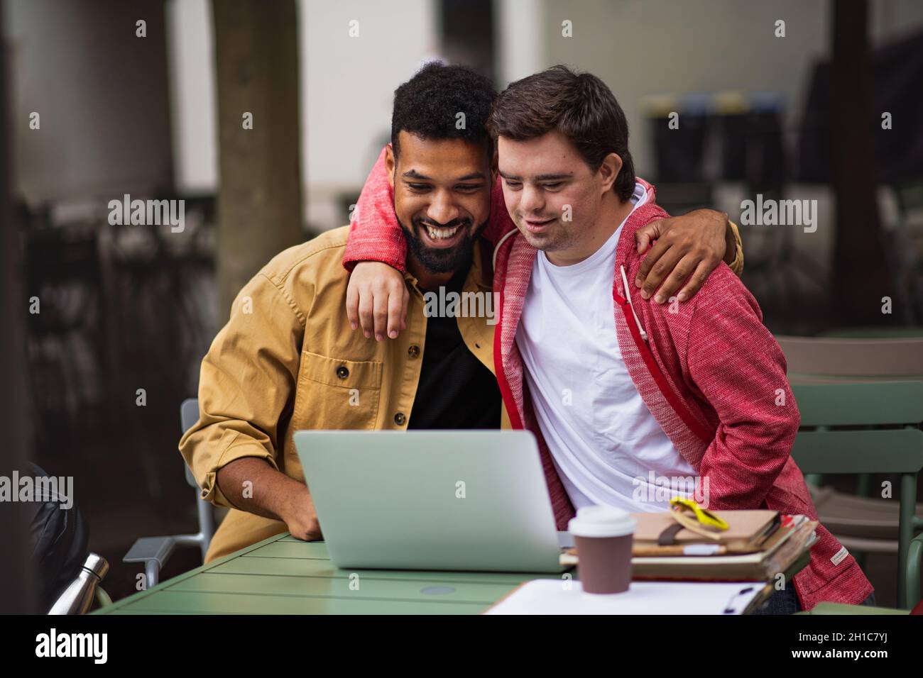 Young man with Down syndrome and mentoring friend with arms around outdoors in cafe using laptop Stock Photo