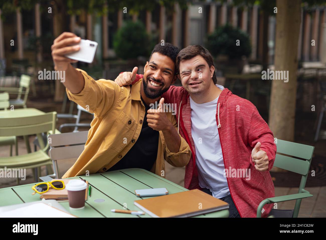 Young man with Down syndrome and his mentoring friend sitting and taking selfie outdoors in cafe Stock Photo