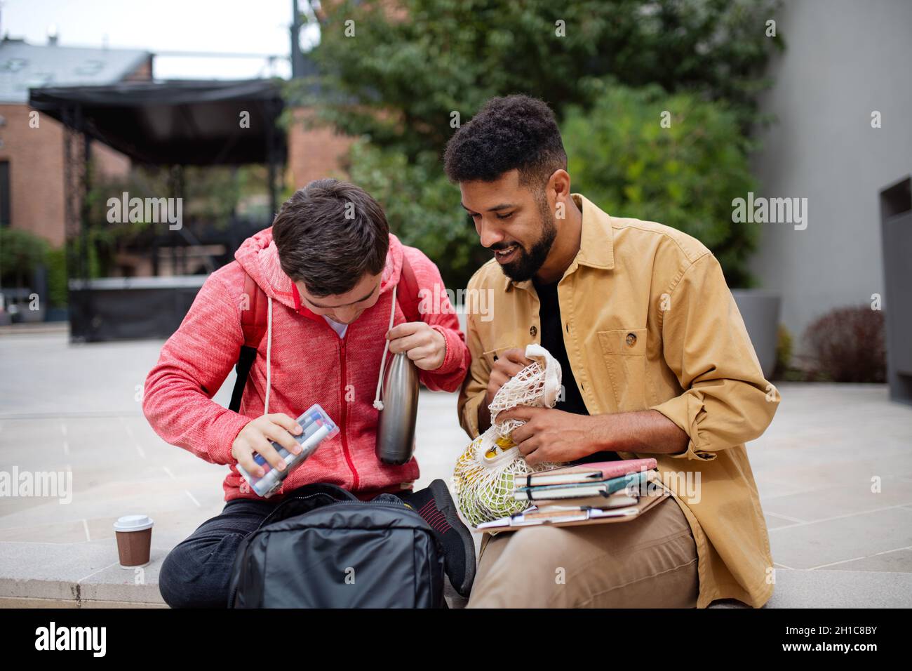 Young man student with Down syndrome and his mentoring friend sitting outdoors in campus area Stock Photo