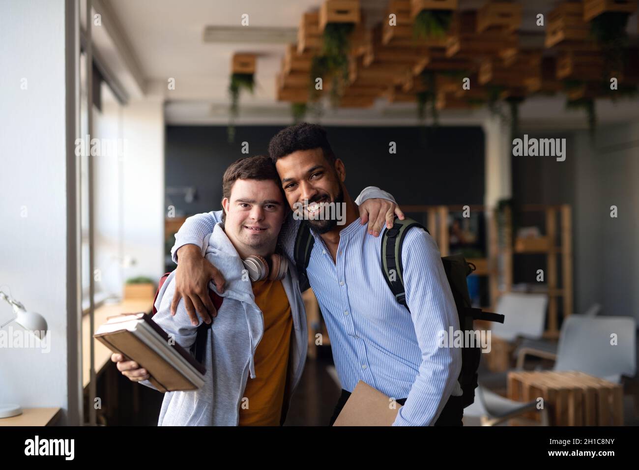 Young man with Down syndrome and his tutor with arms around looking at camera indoors at school Stock Photo