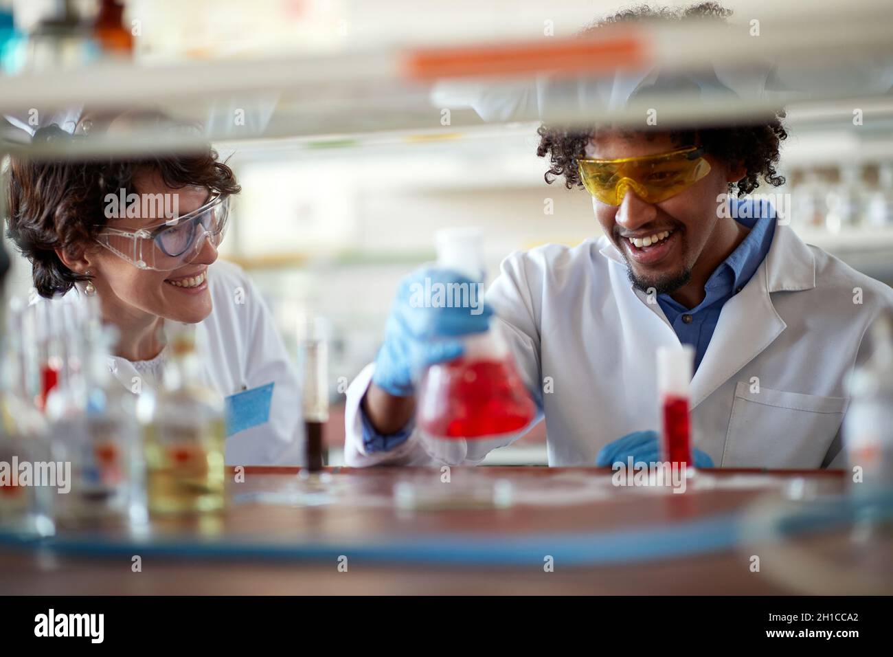 Young colleagues in a protective gear are in a good mood while work with colorful chemicals in a relaxed atmosphere in the university laboratory. Scie Stock Photo