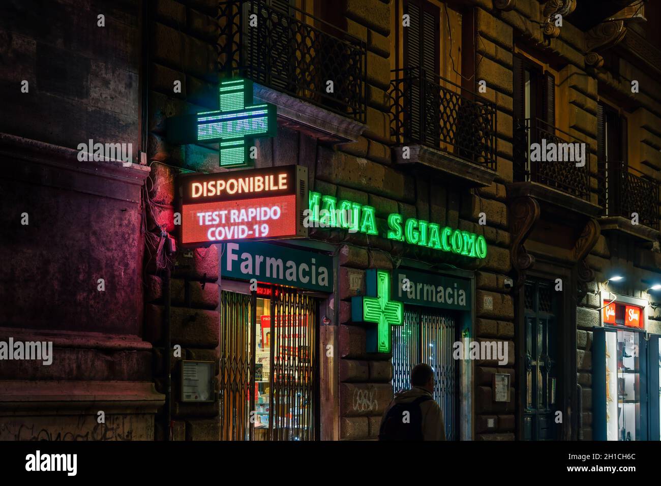 Rome, Italy open pharmacy store entrance with Covid-19 rapid test sign. External night view of Italian pharmaceutical shop with illuminated green display and window showcase. Stock Photo