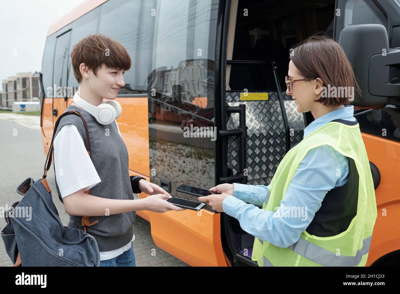 Happy girl passenger holding smartphone under tablet of young female bus conductor checking online ticket Stock Photo