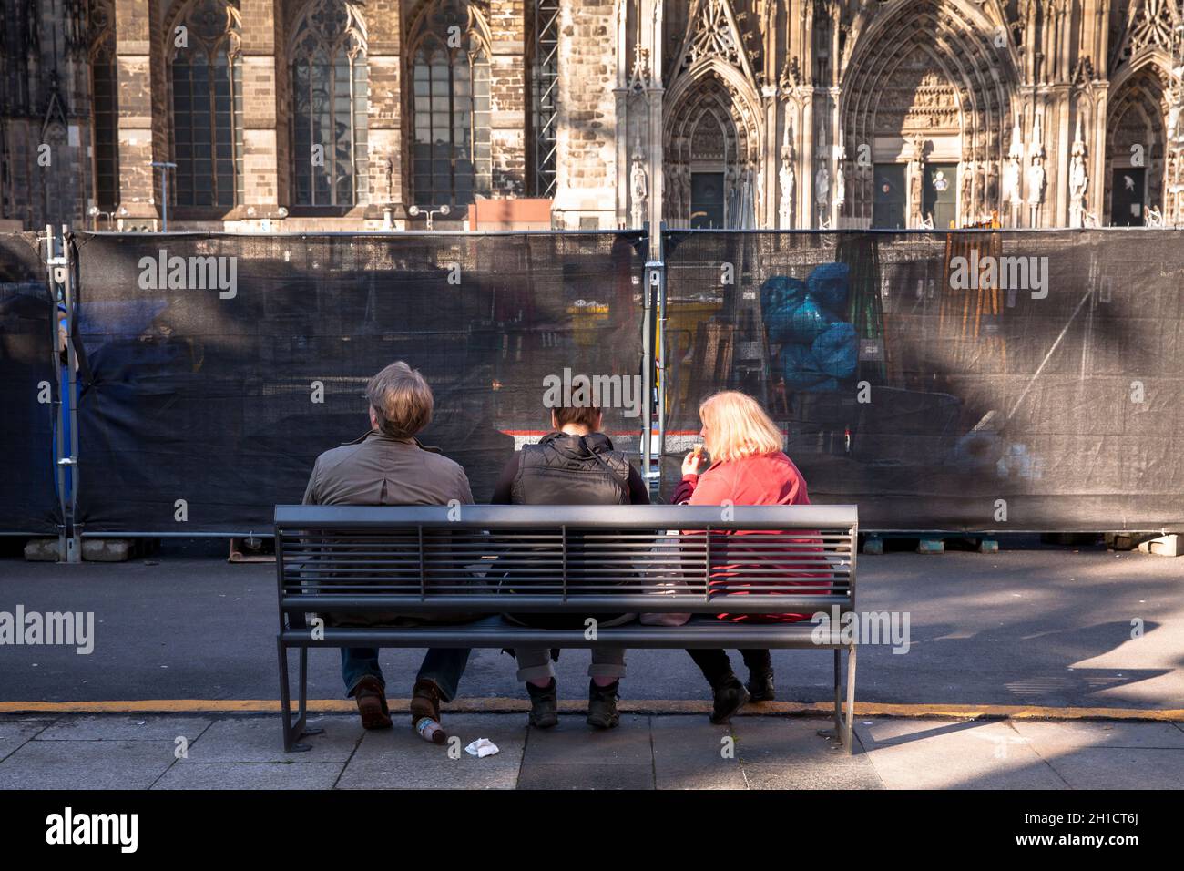 people sitting on a bench in front of a construction fence at Roncalliplatz near the cathedral, Cologne, Germany.  Menschen sitzen auf einer Bank vor Stock Photo