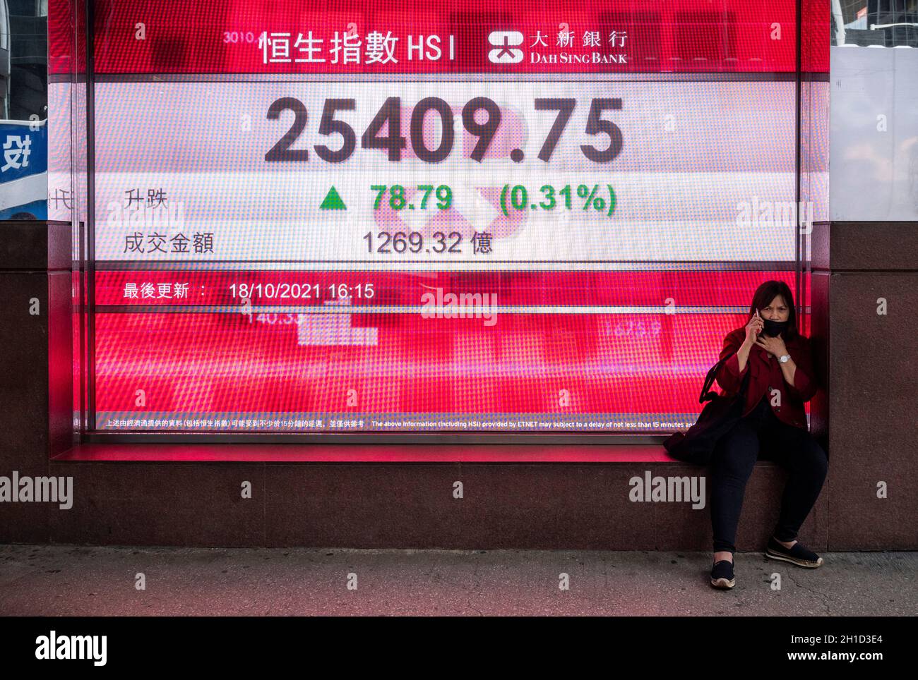 Hong Kong, China. 18th Oct, 2021. A woman sits next to a stock market display board showing the Hang Seng Index information Credit: SOPA Images Limited/Alamy Live News Stock Photo