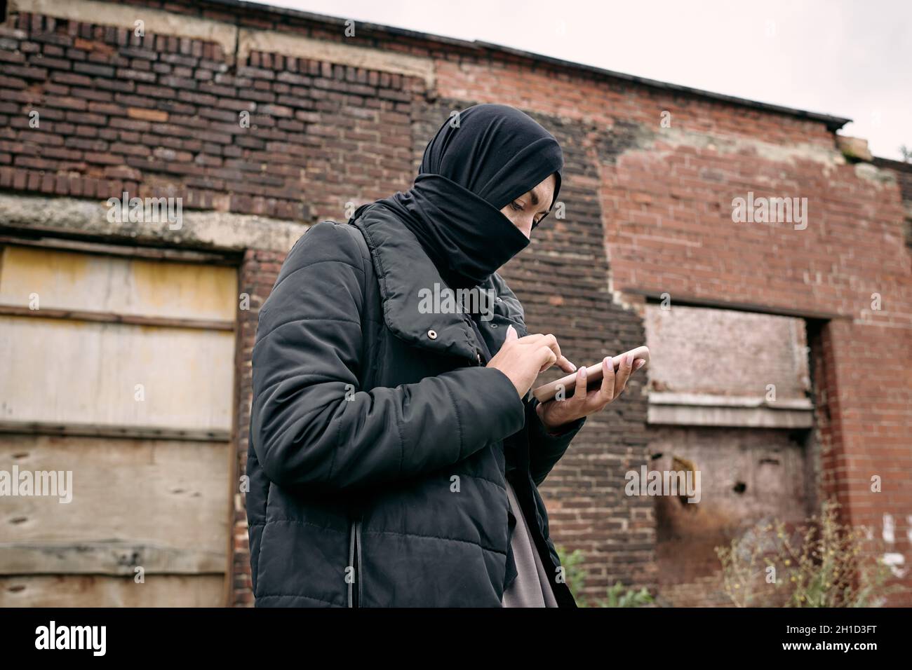 Young refugee female in hijab texting in mobile phone against wall of half ruined building Stock Photo