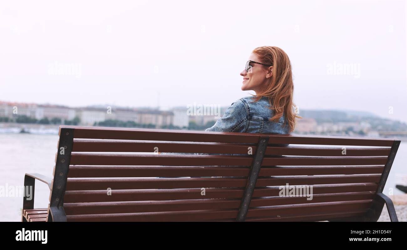 Happy young redhead woman relaxing on bench at riverside in city Stock Photo