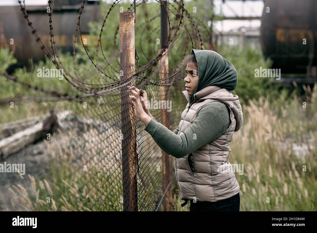Homeless and lonely girl in casualwear standing by barb wire fence on the territory of refugee camp Stock Photo