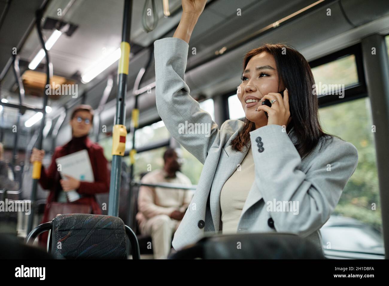 Portrait of smiling Asian woman speaking by smartphone in bus and holding onto handle while traveling by public transport in city, copy space Stock Photo