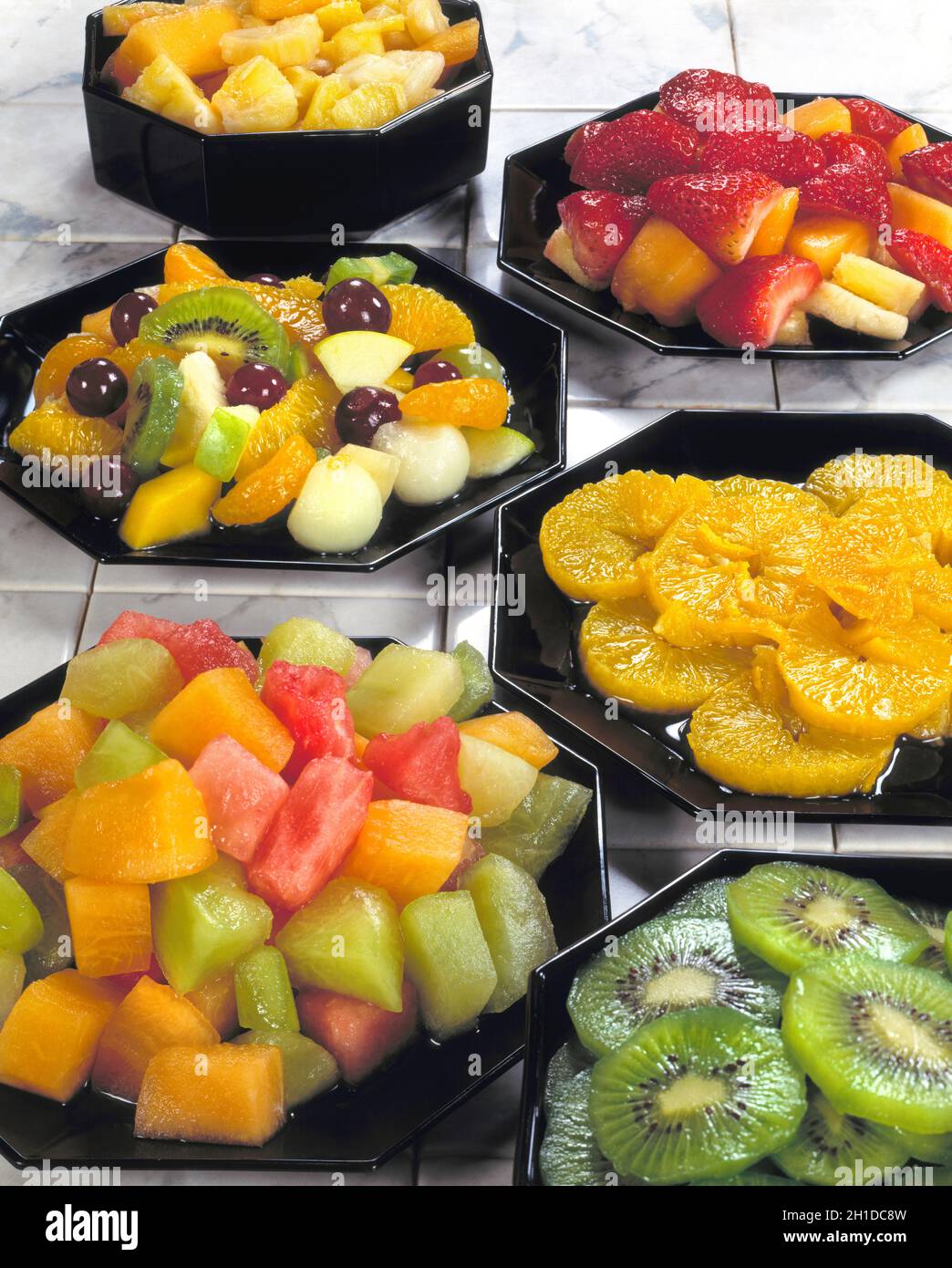 Prepared variety of fruit in black dishes melon cubes oranges kiwi red  strawberries pineapple chunks grapes mango apple white marble tiles background Stock Photo