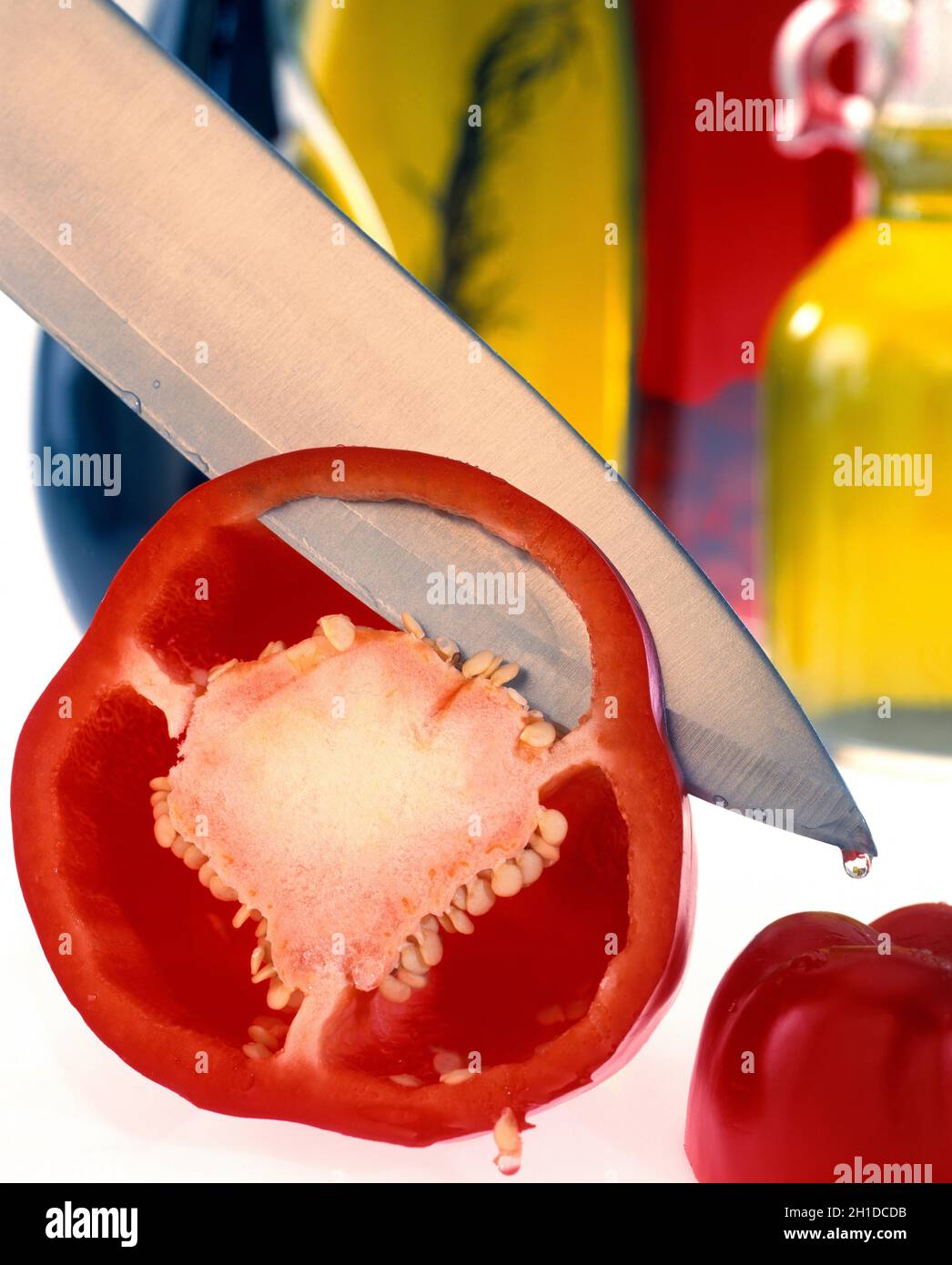 Steel knife cutting slice red pepper on white background soft focus coloured bottles in background Stock Photo