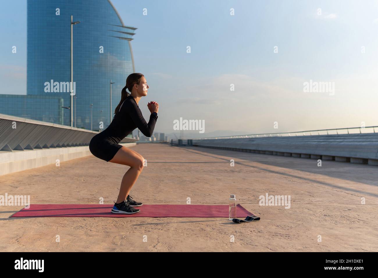 Side view of female athlete in black activewear clasping hands and doing squats on mat during fitness workout early in morning in city. Stock Photo