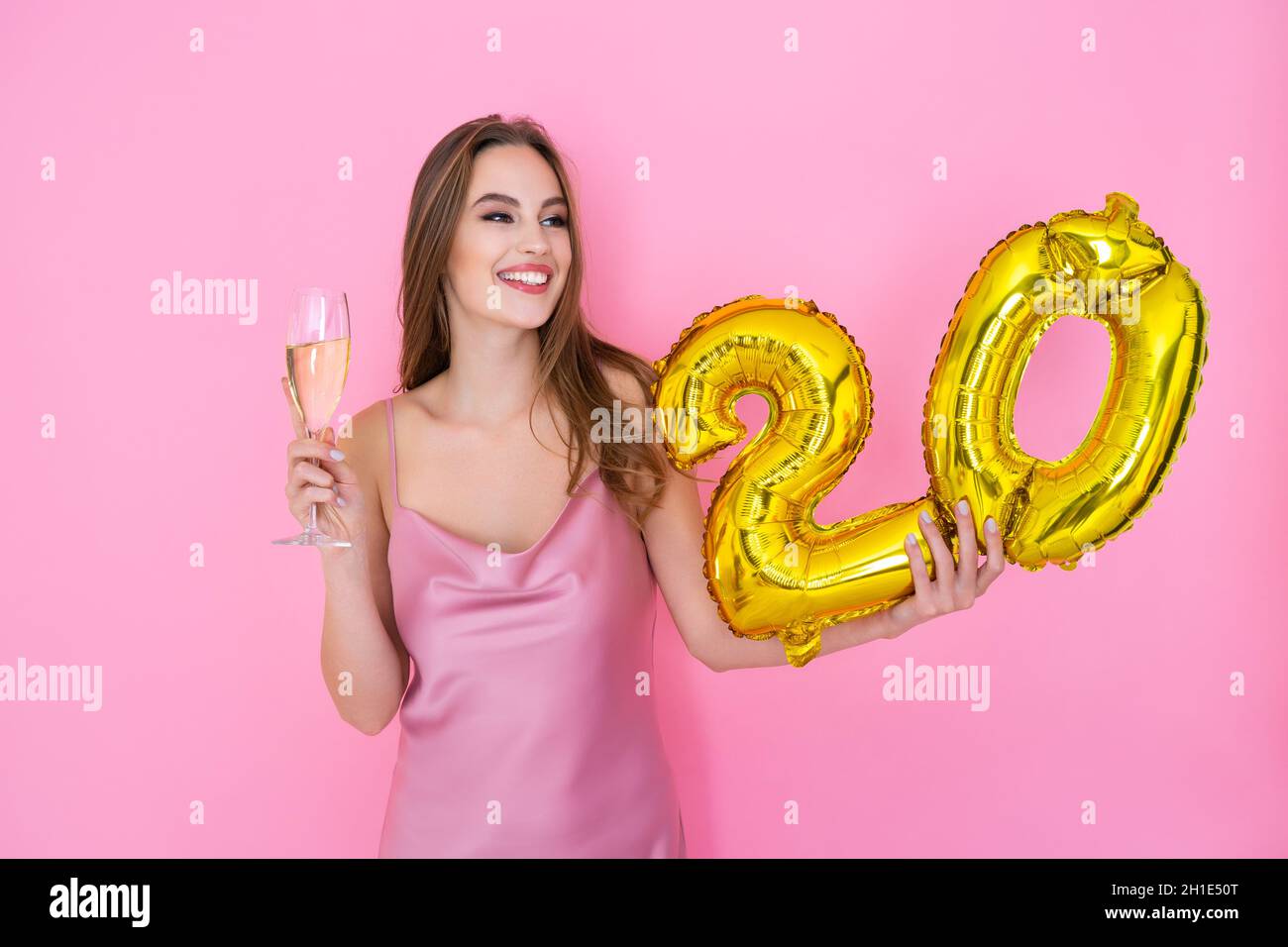 Young happy girl holds gold foil balloon and glass of champagne on pink background. BIRTHDAY PARTY Stock Photo