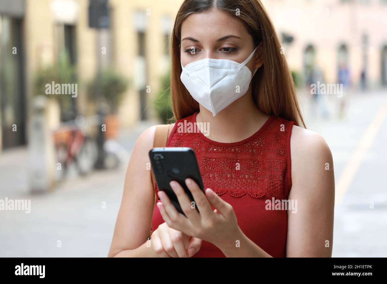 COVID-19 Portrait of Young Woman Wearing KN95 FFP2 Mask Using Smart Phone App in City Street to Aid Contact Tracing and Self Diagnostic in Response to Stock Photo