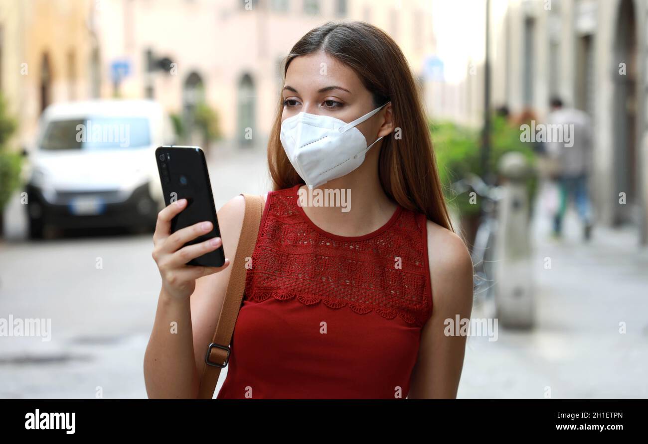 COVID-19 Portrait of Young Woman Wearing KN95 FFP2 Mask Using Smart Phone App in City Street to Aid Contact Tracing and Self Diagnostic in Response to Stock Photo