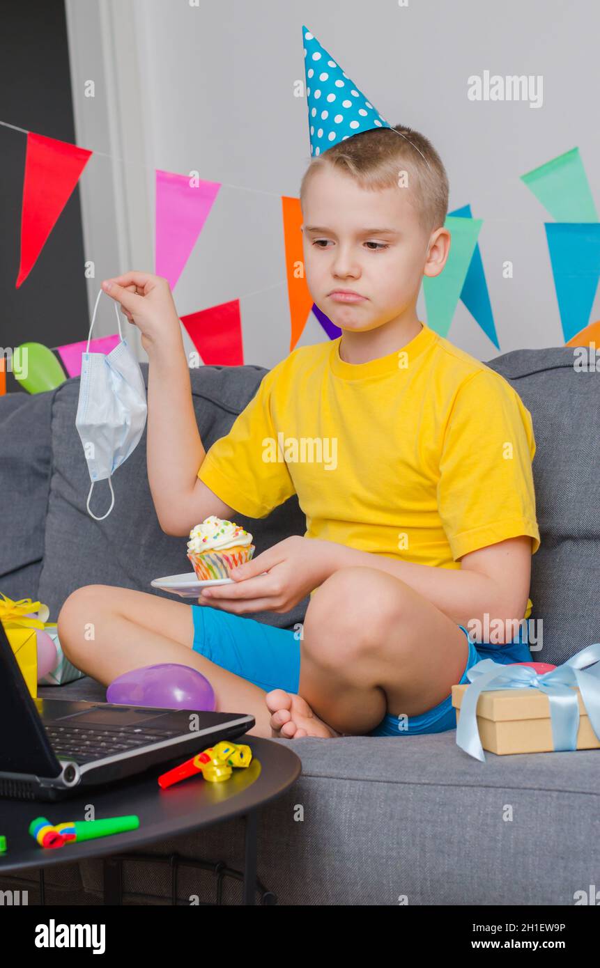 Sad boy with medicine face mask and holiday cupcake celebrates birthday by video call to laptop. Quarantine birthday online in isolation. Virtual even Stock Photo