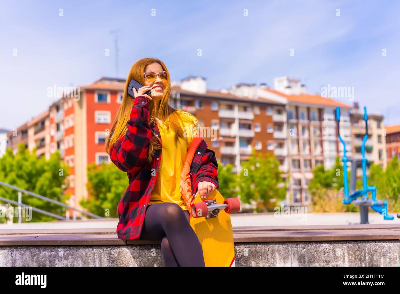 Stylish redhead Hispanic lady talking on her phone in an urban park holding her skateboard Stock Photo