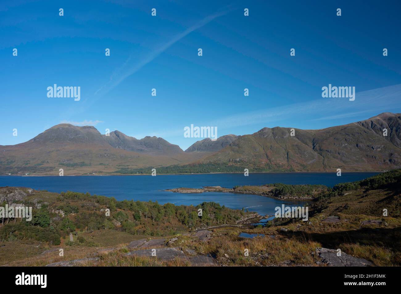 Views over forest and Loch Torridon to Beinn Alligin and Liathach mountains on a sunny day. Blue sky and light cloud with jet stream. No people. Stock Photo