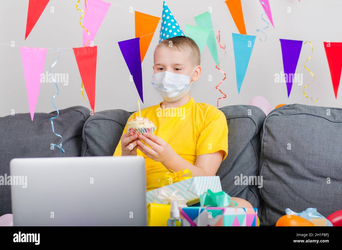 Happy boy in medicine face mask with holiday cupcake celebrates birthday by video call to laptop. Quarantine birthday online in isolation. Virtual eve Stock Photo