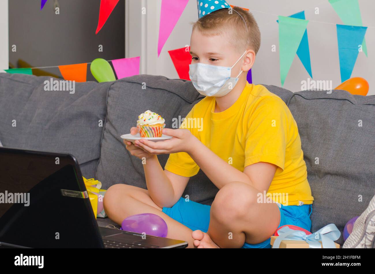 Sad boy in medicine face mask with holiday cupcake celebrates birthday by video call to laptop. Quarantine birthday online in isolation. Virtual event Stock Photo