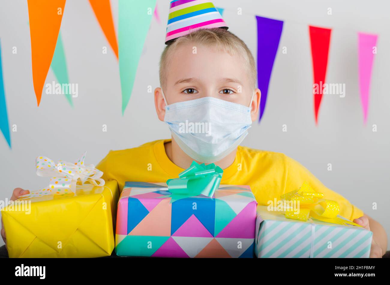 Happy boy in medicine face mask and festive cap with gifts in hand celebrates birthday. Quarantine birthday alone in isolation. Social distance. Stock Photo
