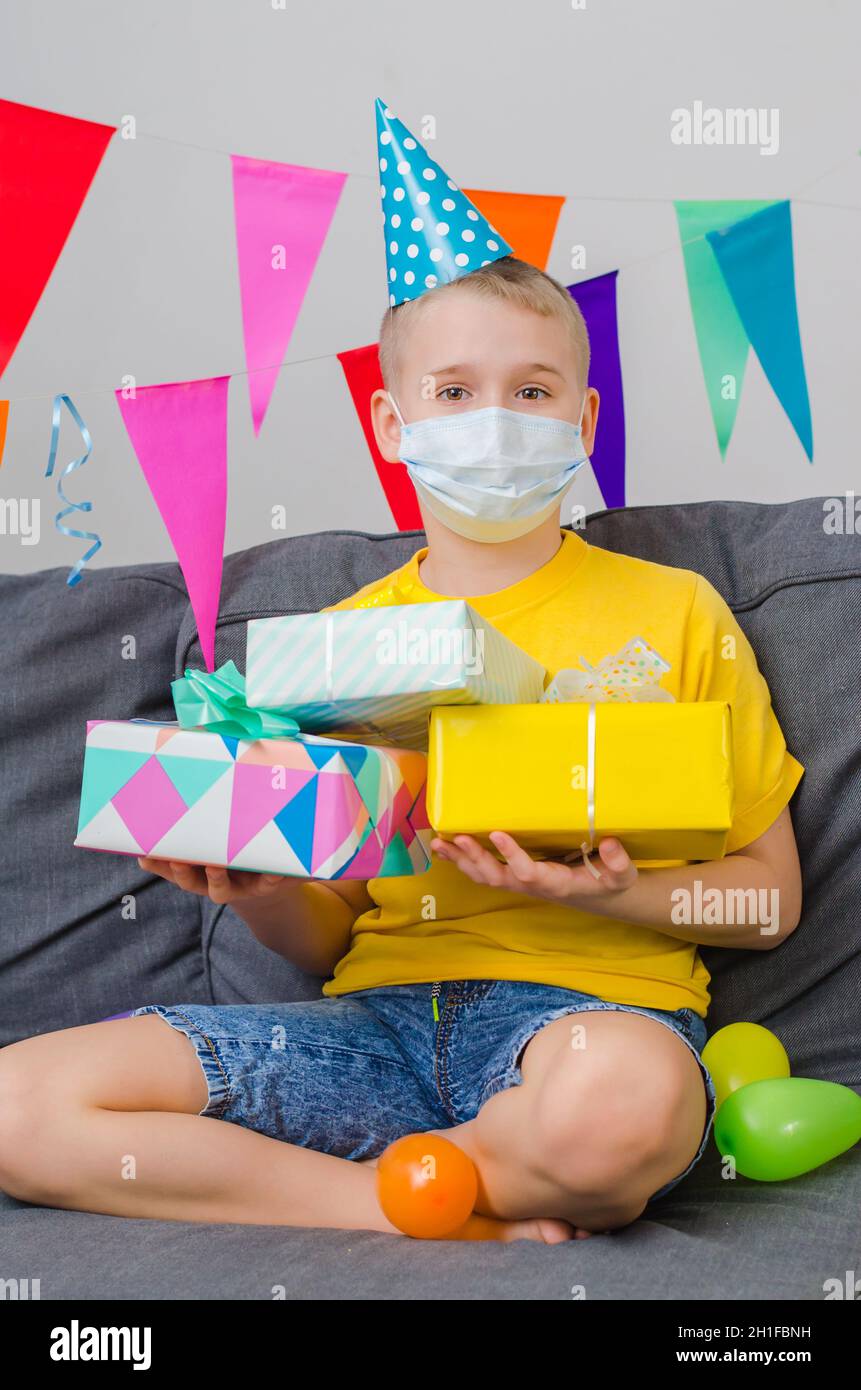 Happy boy in medicine face mask and festive cap with gifts in hand celebrates birthday. Quarantine birthday alone in isolation. Social distance. Stock Photo