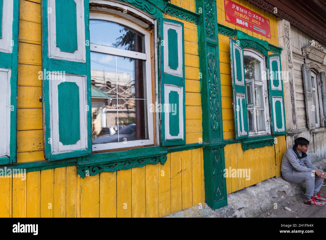 Young man sitting by a green and yellow painted traditional wooden house in use as restaurant in Ulan-Ude. Republic of Buryatia, Russia Stock Photo