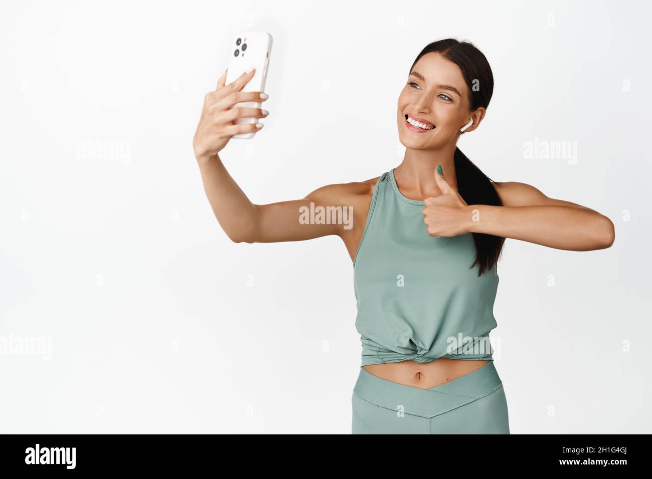 Smiling female fitness instructor record sport video, showing thumbs up  while taking selfie on mobile phone, wearing activewear, white background  Stock Photo - Alamy